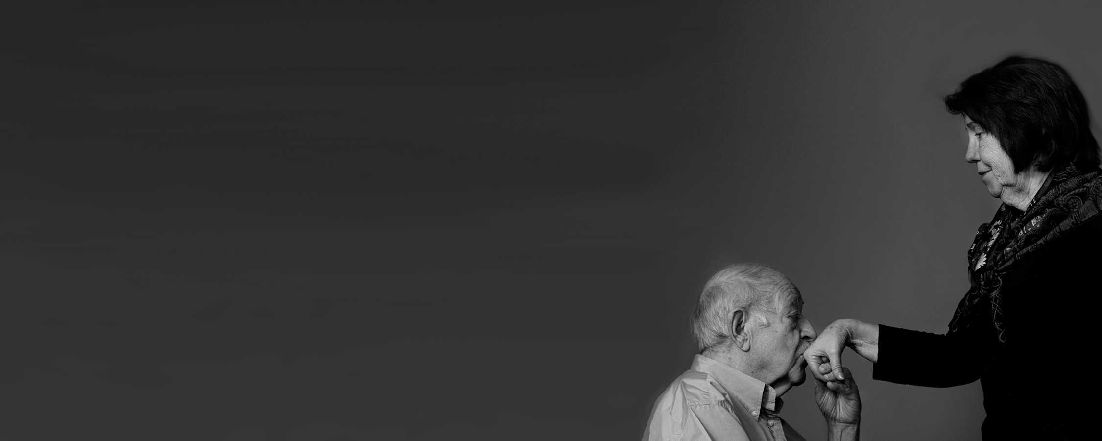 An elderly man kisses the hand of a woman standing beside him